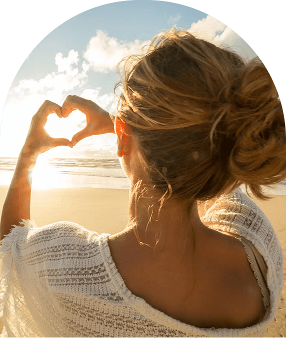Woman making a heart shape with hands at sunset on the beach.